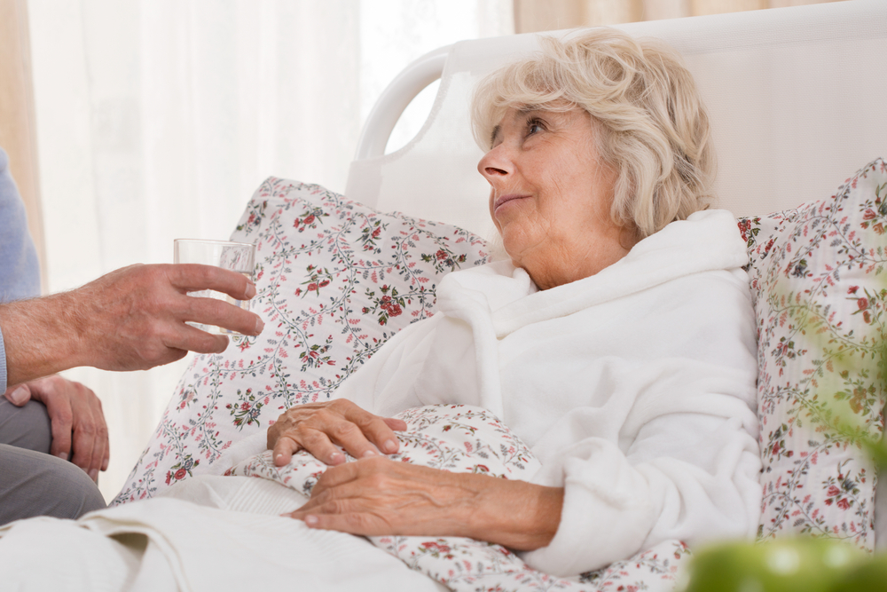 Senior woman sitting upright in bed with someone bringing her glass of water